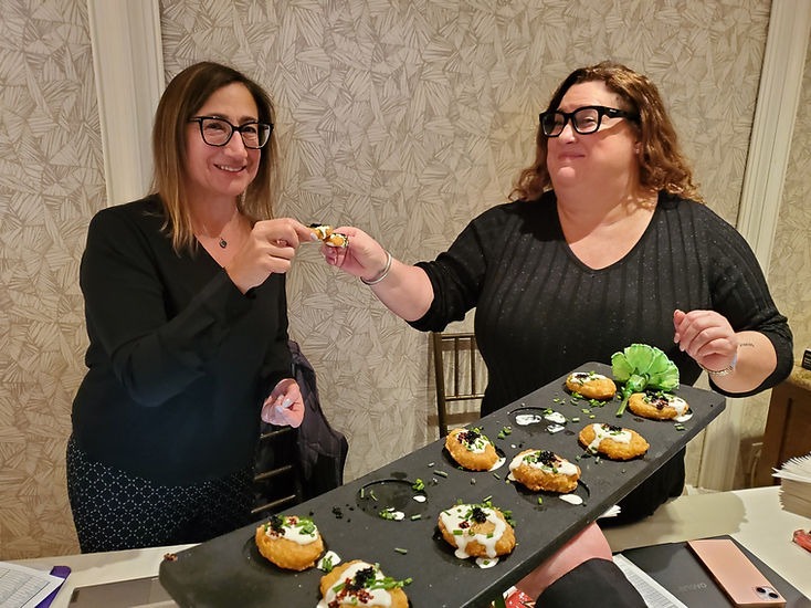 Two women smiling and sharing hors d'oeuvres from a tray in a patterned room. Mood is cheerful. Plates with garnished snacks visible.