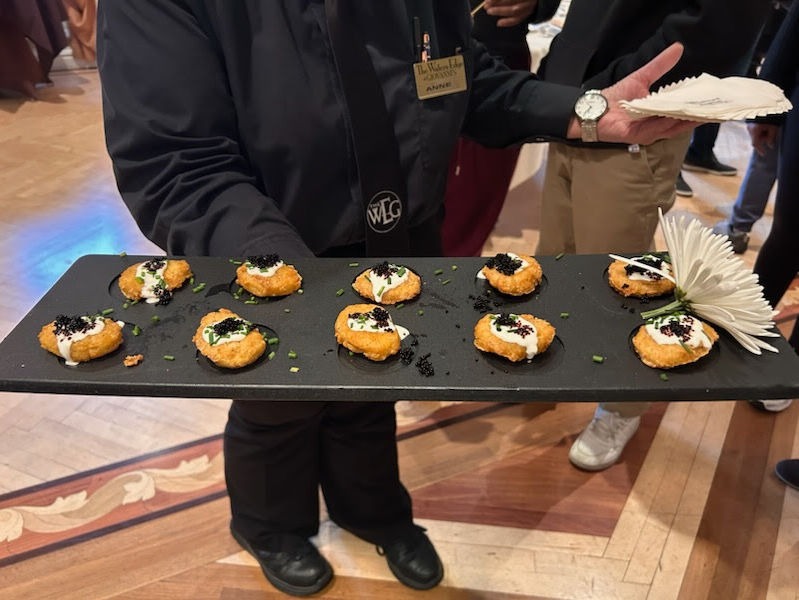 Server in black holds a tray of gourmet appetizers with cream and garnish in an elegant room. A white flower decorates the tray.